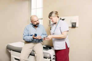 A patient sits up on a medical couch, a nurse standing is demonstrating an insulin pen